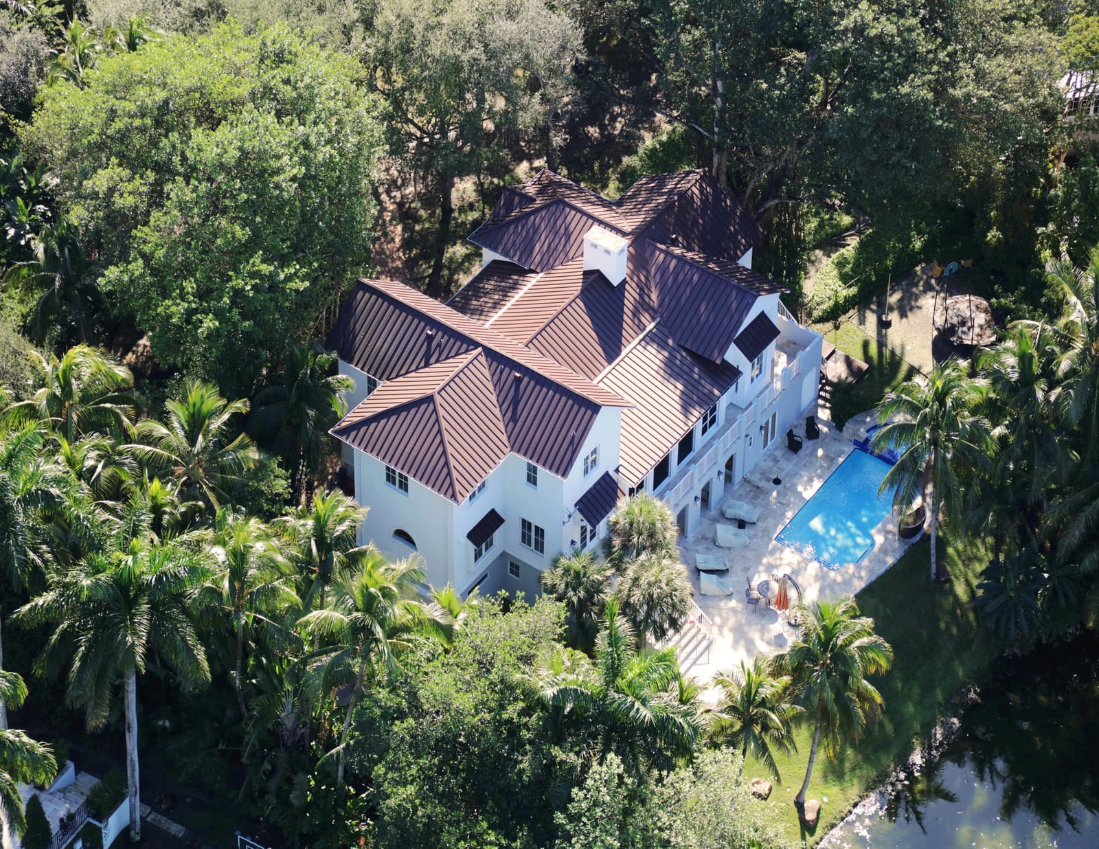 Completed galvalume standing-seam metal roof in a warm bronze finish on a Coral Gables luxury home, aerial three-quarter view amid mature oak and palm canopy