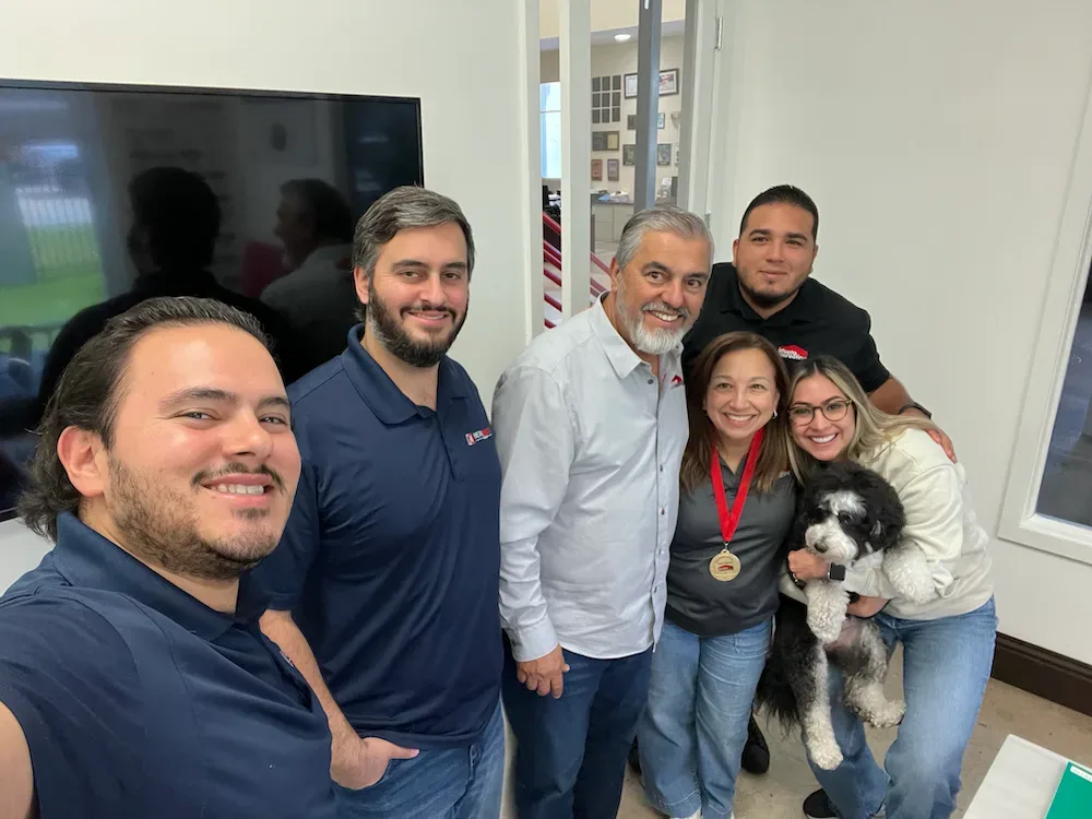 Six members of the Istueta Roofing team smiling together in the office, one holding a small black and white dog